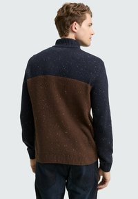 Mann in einem gesprenkelten marineblauen und braunen, gerippten Strickpullover, der leicht seitlich gedreht ist und den Rücken zeigt, kombiniert mit dunklen Hosen.