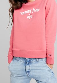 Rosa sweatshirt med vit text "tommy jeans = nyc", ribbad mudd och nederkant; kombinerad med urblekta blå jeans med sidofickor.