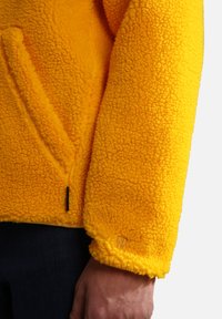 Yellow fleece jacket with a textured, soft surface, featuring a side pocket and a small branded tag at the cuff.