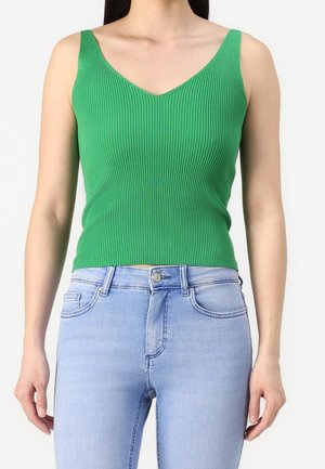 Femme portant un débardeur vert côtelé à col en V assorti à un jean bleu clair, visible des épaules aux hanches sur fond blanc.