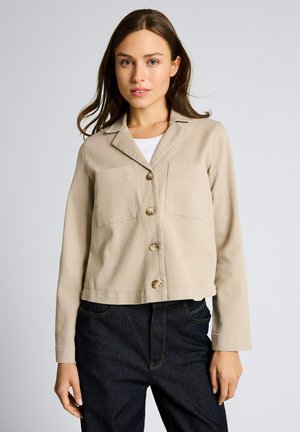 Frau trägt eine beige Jacke mit Knöpfen und Brusttaschen über einem weißen Hemd, kombiniert mit dunkelblauen Jeans, steht vor einem einfarbigen Hintergrund.