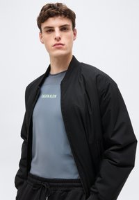 Νεαρός άνδρας που φοράει γαλάζιο πουκάμισο Calvin Klein κάτω από μαύρο μπουφάν με φερμουάρ και μαύρο παντελόνι με κορδόνι, μπροστά σε απλό ανοιχτόχρωμο φόντο.