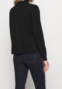 Pull noir à col montant, avec des poignets côtelés et une coupe ample, associé à un jean foncé orné d'un détail à carreaux sur le côté.