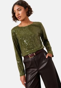 Langärmeliges grünes Paillettentop mit gerippter Textur, kombiniert mit dunkelbraunen Lederhosen und einem kastanienbraunen Gürtel mit goldener Schnalle.