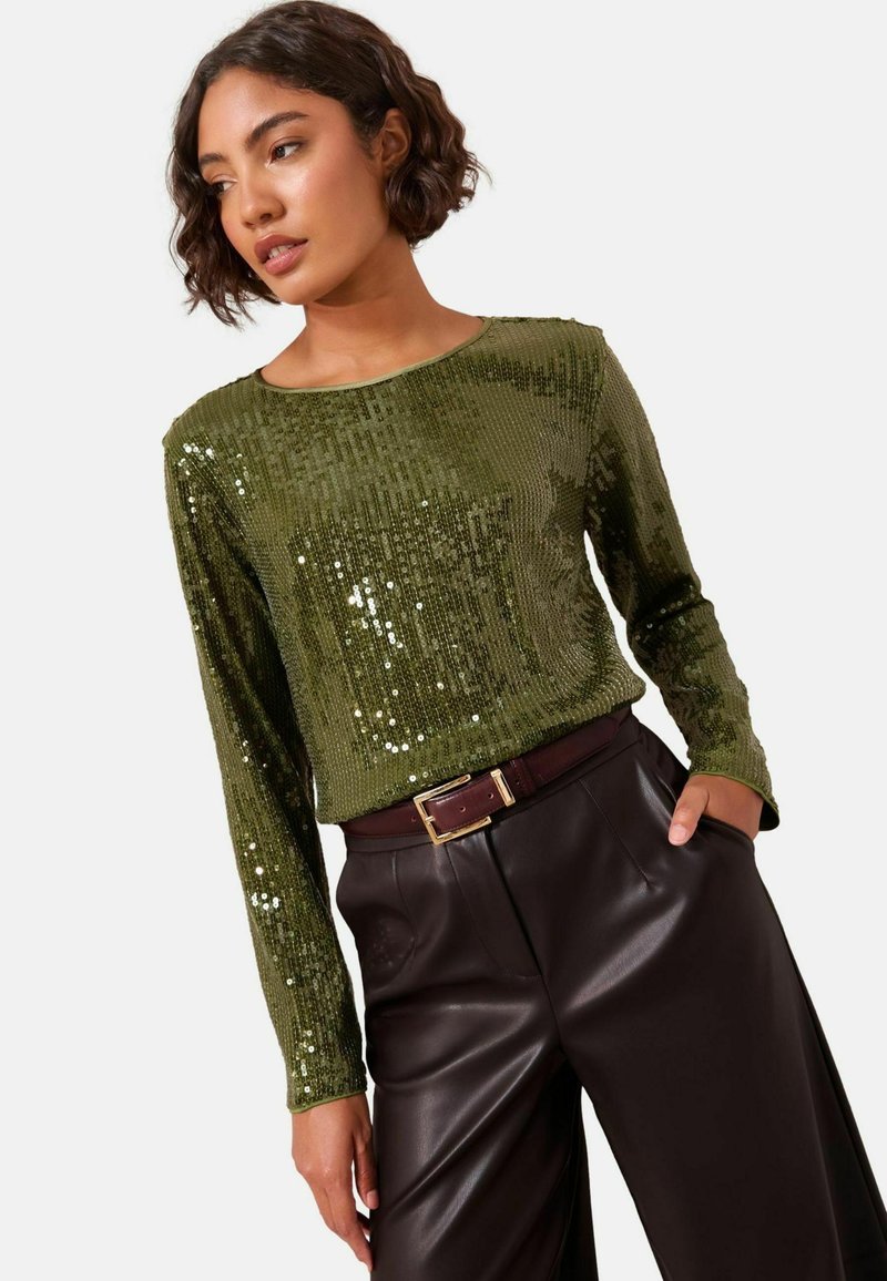 Langärmeliges grünes Paillettentop mit gerippter Textur, kombiniert mit dunkelbraunen Lederhosen und einem kastanienbraunen Gürtel mit goldener Schnalle.