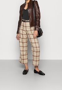 Žena v hnědé kožené bundě, černém topu s krajkovými lemy, béžových kostkovaných kalhotách, s černým opaskem, černými mokasínami a nese černou kabelku přes rameno.