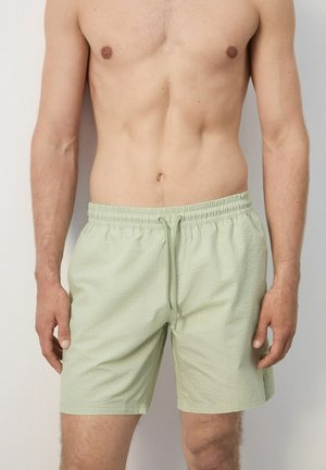 Uomo senza maglietta che indossa pantaloncini verdi chiaro con coulisse e trama, in piedi contro uno sfondo semplice, con le braccia rilassate lungo i fianchi.