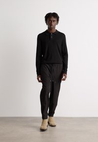 Tricou polo negru cu mâneci lungi și guler, asociat cu pantaloni negri conici. Purtați cu cizme bej până la gleznă, pe un fundal neutru.