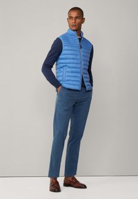 Ljusblå kavad pufferväst med hög krage, tillsammans med en mörkblå långärmad topp och blå byxor. Bruna läder slips-on skor.