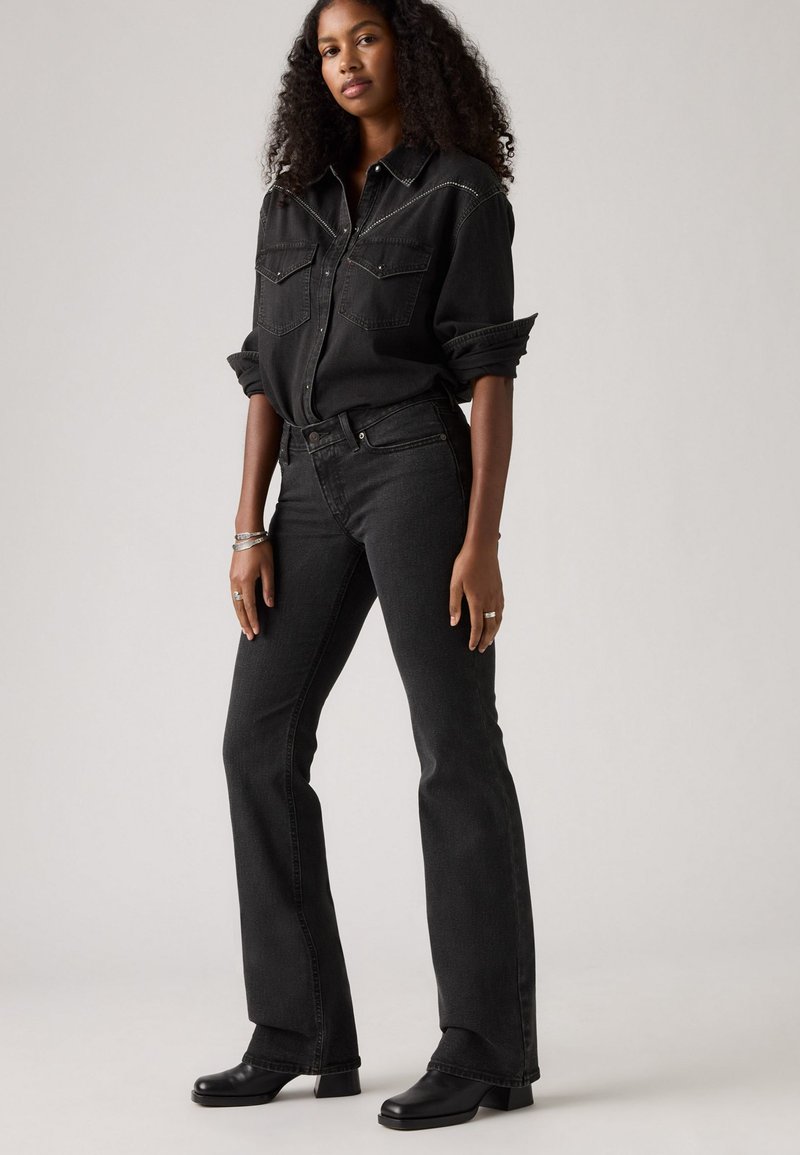 Chemise en denim noir avec boutons-pression, deux poches avant et coutures décoratives, associée à un jean noir évasé et des bottes à talons simples.