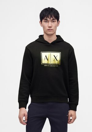 Jonge man met een zwarte hoodie met goudkleurig Armani Exchange-logo, staand tegen een effen lichte achtergrond.
