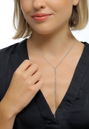 Vrouw die een zilveren Y-vormige ketting draagt met twee hangende kettingen, een zwarte V-hals top en kleine stud oorknopjes, terwijl ze de ketting aanraakt.