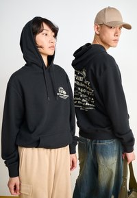 Black hoodies with front pockets, featuring chest and back graphics. One is paired with light beige pants, the other with distressed denim.