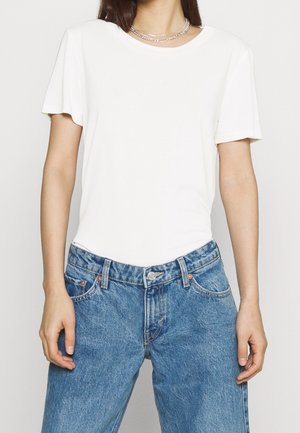 Femme portant un t-shirt blanc uni à manches courtes et un jean bleu taille haute, avec des colliers chaîne en argent.