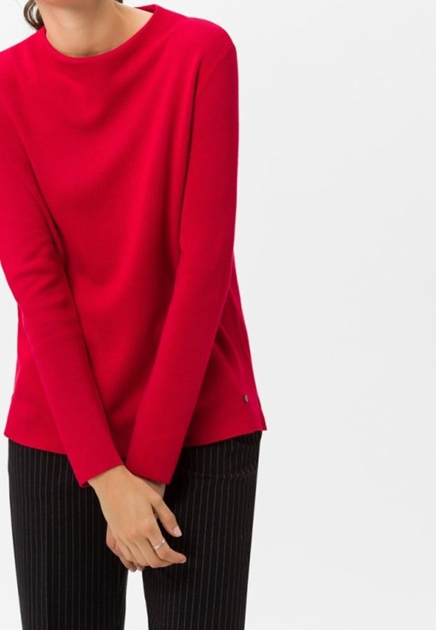 Femme portant un pull rouge vif à manches longues et un pantalon noir à fines rayures, les mains doucement jointes devant.
