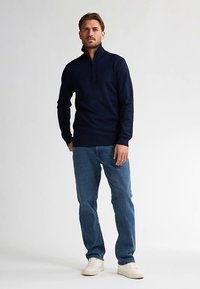 Marineblauwe sweater met een kwartrits van zacht gebreid materiaal, gecombineerd met lichtblauwe jeans en witte sneakers. Eenvoudig ontwerp zonder opvallende patronen.