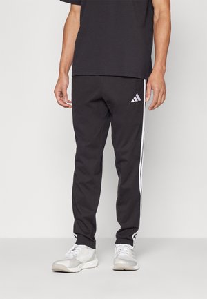 Mann trägt schwarze Adidas-Trainingshose mit weißen Streifen an den Seiten und weiße Sportschuhe, steht auf einem hellgrauen Boden.
