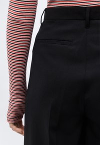 Κοντινό πλάνο μαύρων ψηλόμεσων παντελονιών με πίσω τσέπη welt, φορεμένα με μακρυμάνικο τοπ με κόκκινες, λευκές και μαύρες ρίγες.