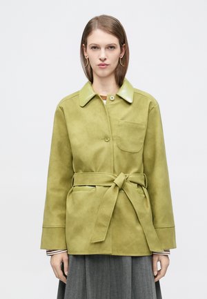 Ženska, oblečena v svetlo zeleno j Jakno z zaponko na ovratniku in žepom na prsih, združeno s sivo plisirano krilo na belem ozadju.