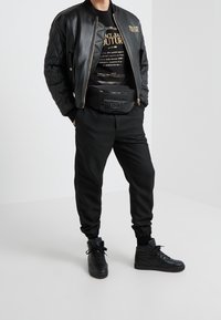 Chaqueta bomber negra con detalle acolchado, acentos dorados, camiseta gráfica negra, joggers negros y zapatillas altas. Riñonera en la cintura.