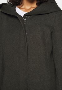 Chaqueta con capucha de color verde oscuro, hecha de un tejido texturizado, que cuenta con un cierre frontal completo con cremallera y botones a presión para mayor seguridad.