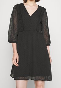 Svart klänning med V-ringning, volangdetaljer och trekvarts genomskinliga ärmar. Har knappdetaljer och ett strukturerat tyg med prickar.