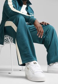 Tyrkysový teplákový souprava s krémovými pruhy a zipovou bundou. Bílé tenisky se zlatými akcenty. Osoba sedí na bílé kovové židli.