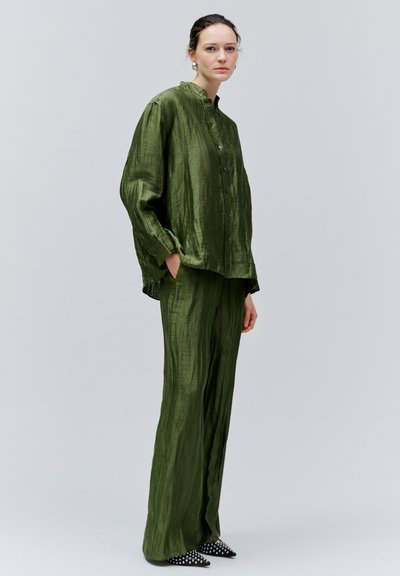 Femme debout portant une chemise boutonnée ample texturée vert olive et un pantalon large assorti avec des chaussures noires pointues à pois blancs.