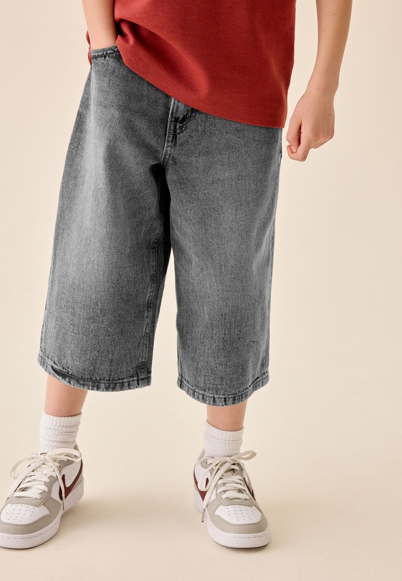 Niño con pantalones cortos de mezclilla grises hasta la rodilla, calcetines blancos y zapatillas blancas con detalles en rojo, de pie sobre un fondo claro.