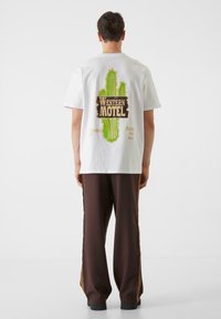 Bijela kotonska majica s dizajnom zelenog kaktusa i tekstom "WESTERN MOTEL" na leđima. Uparen s smeškim širokim hlačama.