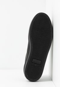 Semelle de basket noire avec motif de bande de roulement en zigzag et "NO NAME" en relief au centre, positionnée à la verticale contre un coin blanc.