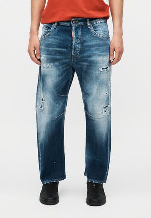 Mann trägt abgenutzte blaue Jeans und schwarze Sneaker, steht mit entspannten Händen an den Seiten vor einem einfarbigen Hintergrund.