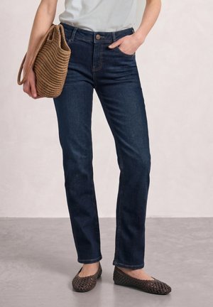 Femme portant un jean droit bleu foncé, des ballerines tissées marron, tenant un sac à main tissé marron et portant un haut blanc.
