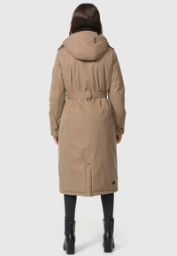 Lange beige jas met een capuchon, getailleerde taille en een achterventilatie. Heeft een gestructureerde stof en zijzakken, gecombineerd met zwarte legging en laarzen.