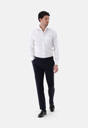 Camicia bianca con bottoni e tasca sul petto, abbinata a pantaloni navy e scarpe nere. La camicia ha una texture liscia e una vestibilità su misura.