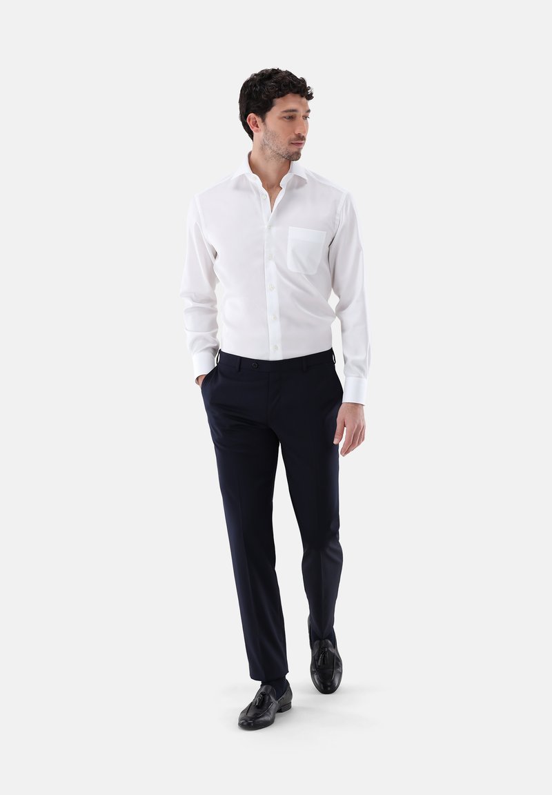 Weißes Hemd mit Knöpfen und einer Brusttasche, kombiniert mit navyblauen Hosen und schwarzen Schuhen. Das Hemd hat eine glatte Textur und eine taillierte Passform.