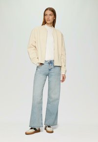 Lichtbeige gewatteerde bomberjack, witte turtleneck, lichtblauwe wijde jeans en casual sneakers. Het jack heeft ribbroekmanchet en onderrand.