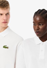 Białe koszulki polo z zielonym wyszywanym logo krokodyla. Materiał wygląda na teksturowany, z klasycznym kołnierzykiem i zapięciem na guziki.