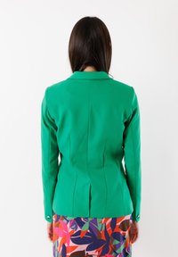 Blazer verde su misura con taglio aderente, colletto a scolla e dettagli delle cuciture posteriori. Maniche rifinite con accenti di bottoni argento.