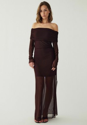 Femme portant une robe marron foncé épaules dénudées avec de longues manches transparentes et une longue jupe transparente, debout devant un fond uni.