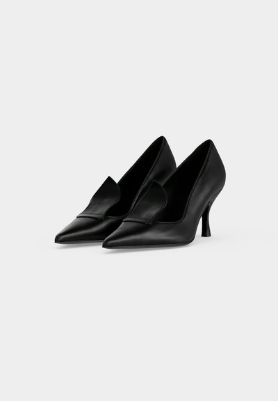 Paire de chaussures à talons hauts noires à bout pointu avec un talon bas et fin sur fond blanc.