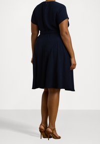Navy blue knee-length dress with short sleeves, fitted waist, and flared skirt. Smooth texture with a back zipper. Worn with brown high-heeled sandals.