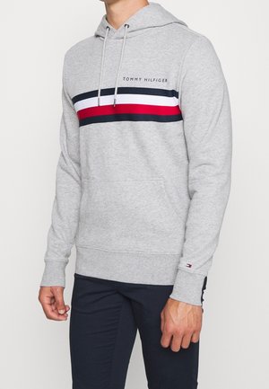 Mężczyzna ubrany w jasnoszarą bluzę z kapturem Tommy Hilfiger z granatowymi, białymi i czerwonymi poziomymi paskami oraz granatowe spodnie na jednolitym tle.