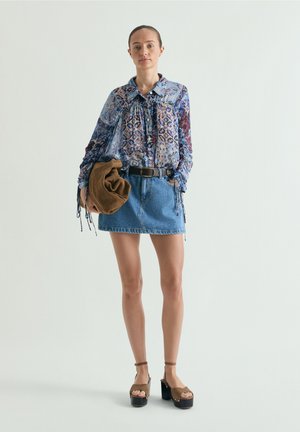 Mujer de pie, vistiendo una blusa de manga larga con estampado, minifalda de mezclilla, sandalias con plataforma y sosteniendo un bolso marrón doblado en una mano.