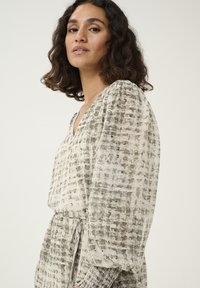 Blouse à manches longues et décolleté en V en tissu léger à motif, présentant un design en quadrillage dans des tons beiges et gris atténués, avec des manches froncées.