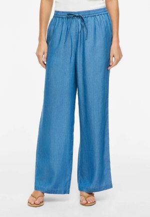 Kvinne iført løse lyseblå denim-bukser med elastisk midjebånd og snøring, kombinert med brune flip-flop-sandaler.