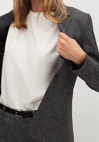 Blazer à carreaux gris avec un design structuré, fermeture à un bouton et doublure noire, porté sur une chemise blanche, associé à une ceinture noire.