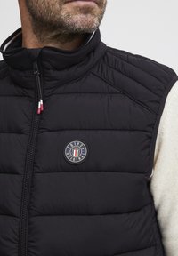 Svart puffer-vest med høy krage, utstyrt med en sirkulær logo-patch, glidelås og horisontal søm for tekstur.