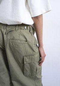 Άτομο που φοράει χαλαρό χακί cargo παντελόνι με  πλαϊνές τσέπες και λευκό κοντομάνικο πουκάμισο, στέκεται μπροστά σε φωτεινό φόντο.