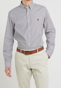 Skjorta med knäppning i marinblå och vita vertikala ränder, med en spetsig krage, långa ärmar och en liten broderad logotyp på bröstet.